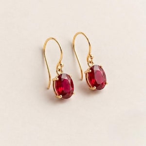 Puede incluir: Un par de pendientes colgantes con piedras preciosas de color rojo intenso en forma ovalada engastadas en metal dorado. Cada pendiente cuelga de un gancho de alambre de oro curvado, mostrando una piedra facetada sujeta por cuatro garras.