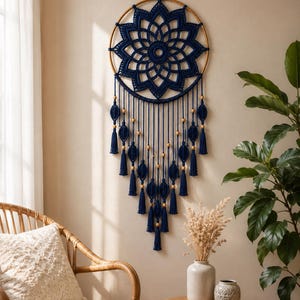 Peut inclure: Une décoration murale composée d'un cercle en bois orné d'un motif mandala en corde macramé bleu marine foncé. Le design comprend des fils suspendus agrémentés de perles en bois clair et de pompons bleu marine disposés en forme de V.