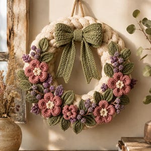 Può includere: Una ghirlanda circolare realizzata in spesso filato intrecciato color crema, caratterizzata da un grande fiocco all'uncinetto verde oliva sulla parte superiore. La ghirlanda è decorata con fiori rosa all'uncinetto con centri crema, piccoli grappoli di bacche viola e foglie verdi.