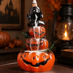 May include: A ceramic incense burner featuring a stack of three orange jack-o'-lanterns topped with a small black ghost figure. A cone-shaped incense sits atop the ghost, releasing white smoke that cascades down the pumpkins like a waterfall.