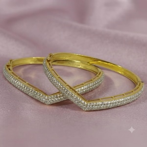 May include: Two gold-toned hinged bangles featuring a V-shaped silhouette. Each bangle is encrusted with numerous small, clear, sparkling stones along the outer surface, creating a continuous pavé effect across the metal frame.