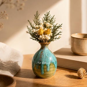 May include: A small ceramic vase with a rounded body and a narrow neck, featuring a turquoise and olive green drip glaze finish. The text "To Ethan" is printed in white on the front. It holds a bouquet of dried greenery, white baby's breath, and yellow roses.