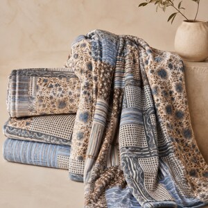 May include: A folded quilt featuring a patchwork design with various patterns including blue and brown floral motifs, geometric grids, and vertical stripes. The fabric displays a mix of muted tones, creating a layered appearance with soft textures.