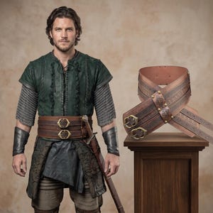 May include: A wide brown leather belt featuring intricate embossed patterns, two brass buckles, and a central decorative plate. The belt is displayed on a wooden pedestal next to a person wearing a matching belt, a green tunic, chainmail sleeves, and black leather bracers.