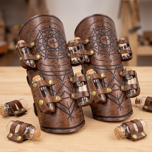 May include: Two brown leather bracers featuring black geometric star patterns and multiple attached small glass vials with cork stoppers. Each bracer has leather loops secured with brass-colored metal rivets to hold the clear glass bottles.