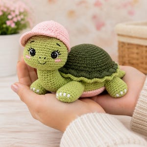 May include: A crocheted turtle toy featuring a light green head and body, a dark green shell, and a light pink baseball cap. The turtle has large black eyes with long eyelashes, a small stitched smile, and pink blush marks on its cheeks.