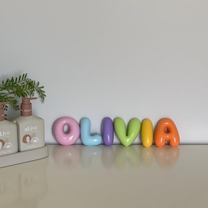 May include: A set of six individual, smooth, balloon-style letters spelling the name OLIVIA in a vibrant colour palette. The letters are pink, light blue, purple, lime green, yellow, and orange, arranged in a horizontal line on a flat surface.