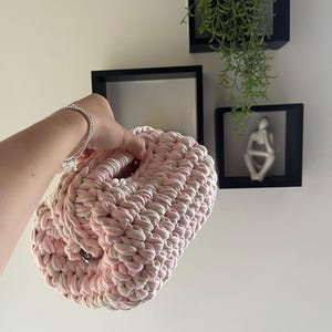May include: A cylindrical crochet bag crafted from thick, soft yarn in a variegated blend of pale pink and off-white tones. The bag features a dense, textured stitch pattern with a circular opening at the top and a sturdy, rounded structure.