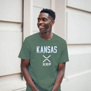 Puede incluir: Una camiseta de manga corta en color verde salvia con la palabra KANSAS en letras mayúsculas blancas sobre un logotipo de criptomoneda XRP blanco y el texto XRP centrado en el pecho. La camiseta tiene cuello redondo y corte estándar.