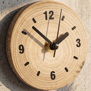 May include: A circular wall clock crafted from a natural wood slice featuring visible grain patterns. The clock face displays black numerals at the 12, 3, 6, and 9 positions, with black hour and minute hands and a central gold-toned metal mechanism.