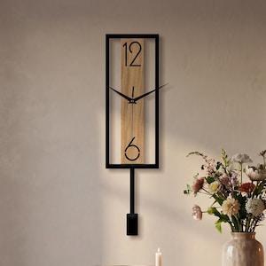 Puede incluir: Un reloj de pared rectangular con un marco de metal negro que rodea un panel vertical de madera clara. La esfera muestra los números negros 12 y 6, con manecillas negras delgadas y un péndulo negro que cuelga debajo del marco.