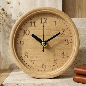 May include: A round wooden table clock with a light brown natural wood grain finish. The clock face features black Arabic numerals from 1 to 12, black hour and minute hands, and a thin black second hand. The clock has a smooth, circular frame.