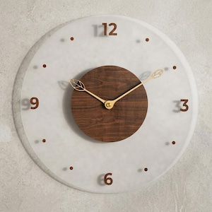 May include: A round wall clock featuring a frosted white glass face with brown numerals at 12, 3, 6, and 9 o'clock. The center consists of a circular dark wood panel with gold-colored metal hands shaped like leaves at the tips.