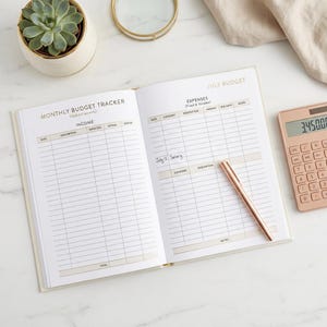 Peut inclure: Un planificateur de budget mensuel ouvert avec des colonnes pour les revenus et les dépenses, affichant le texte July Budget, Monthly Budget Tracker et Expenses. Un stylo or rose est posé sur la page et une calculatrice or rose affiche 3450.00.