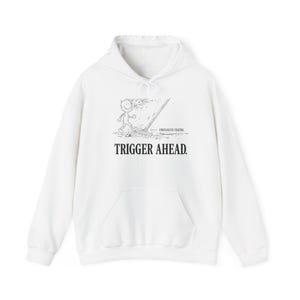 Può includere: Una felpa con cappuccio bianca con una stampa grafica nera sul petto. Il design raffigura un personaggio dei cartoni animati che cammina con un rastrello, accompagnato dal testo "UNRESOLVED TRAUMA" e dal testo "TRIGGER AHEAD." in grassetto.