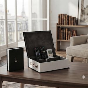 May include: A rectangular storage box designed to look like a book with the word PARIS printed on the side, featuring an open lid and interior compartments holding three remote controls, accompanied by a smaller matching black book-shaped box.