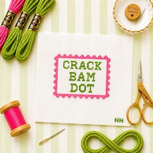 May include: A white cross-stitch fabric square featuring the green text CRACK BAM DOT enclosed in a pink scalloped border. The set includes three green embroidery floss skeins, one pink spool, gold scissors, a needle, and a wooden needle minder.