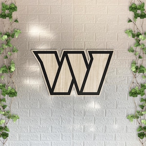 May include: A wooden wall sign featuring the letter W in a bold, geometric font. The letter has a light wood grain finish with a thick, solid black border outlining its shape, creating a sharp contrast against the light-coloured surface.