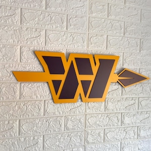 May include: A wall-mounted sign featuring a large, stylised letter W in dark burgundy with a bright yellow border. An arrow shape in the same yellow and burgundy colour scheme extends horizontally through the centre of the letter W, pointing right.