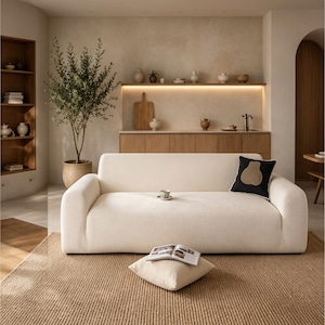 May include: A cream-colored, low-profile sofa with rounded arms and a textured fabric finish. A single black throw pillow with a tan pear-shaped graphic sits on the right side, while a small ceramic cup rests in the center of the seat cushion.
