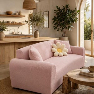 May include: A light pink sofa with a textured herringbone pattern, featuring two matching side cushions and one decorative white and yellow flower-shaped pillow. The sofa has a structured frame with rounded armrests and a solid, uniform fabric finish.