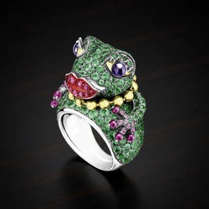 May include: A ring shaped like a frog, featuring a pavé setting of green stones covering the head and body. The frog has prominent red stone lips, purple stone eyes, a gold bead necklace detail, and pink stone accents on the feet and hands.