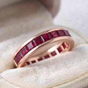 Può includere: Una fede a giro in oro rosa con un incastonatura a binario continua di rubini rosso scuro a taglio quadrato. L'anello presenta una finitura in metallo lucido e una larghezza uniforme con una serie di pietre preziose sfaccettate.