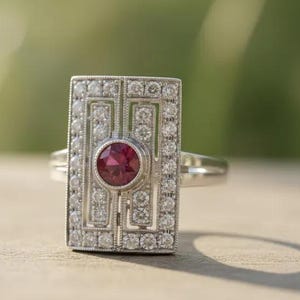 May include: A rectangular silver ring featuring a central round red gemstone surrounded by a geometric frame of small clear stones. The metal surface has a milgrain texture along the edges, and the band is a simple polished silver metal.