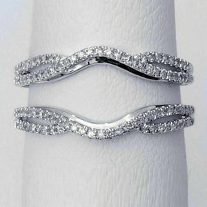 May include: A silver-toned ring guard featuring two curved bands that meet in a central point. Each band is encrusted with small, clear, sparkling stones along the entire length, creating a continuous shimmering effect across the metal surface.