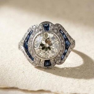 May include: A silver-toned ring featuring a large, round, faceted centre stone surrounded by a halo of small, clear, round stones. The band and setting are accented with deep blue, rectangular-cut sapphires and additional small, clear stones.