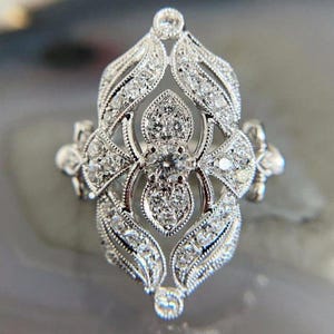 Può includere: Un anello color argento con una parte frontale allungata a forma di marquise e un'intricata lavorazione in filigrana. Il design è incastonato con numerose piccole pietre trasparenti a taglio rotondo, disposte in un motivo simmetrico a petalo attorno a una pietra centrale.