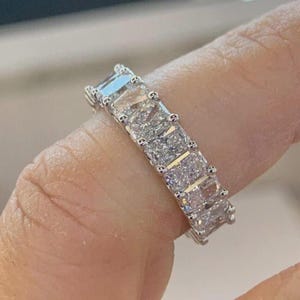 May include: A silver-toned eternity band ring featuring a continuous row of rectangular, radiant-cut clear stones set in a shared-prong metal setting that encircles the finger, showcasing high-clarity faceted surfaces that reflect light.