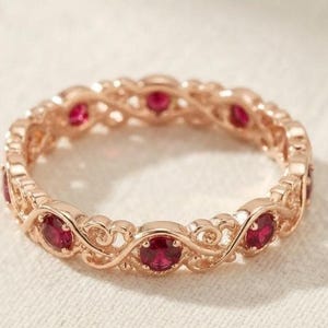 Puede incluir: Una alianza de oro rosa con una serie de rubíes rojos redondos y vibrantes engastados en un intrincado patrón de filigrana metálica entrelazada que envuelve continuamente toda la circunferencia del anillo circular.