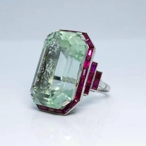 May include: A ring featuring a large, rectangular, light green gemstone set in a metal frame. The central stone is surrounded by a border of smaller, rectangular, deep red rubies arranged in a stepped, geometric pattern along the sides.