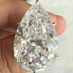 May include: A large, clear, pear-shaped faceted gemstone set in a metallic prong setting. The stone features numerous triangular and kite-shaped facets that reflect light, creating a brilliant, sparkling appearance with sharp, defined edges.