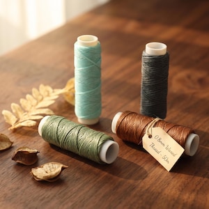 Può includere: Quattro rocchetti di filo cerato in azzurro, grigio scuro, verde salvia e marrone. Il rocchetto marrone presenta una piccola etichetta di carta legata con uno spago che riporta la scritta 0.8mm Waxed Thread - 50m. Ogni rocchetto ha un nucleo bianco.