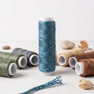 May include: A vertical spool of teal waxed thread stands in the center, surrounded by several other spools in shades of olive green, gray, beige, and brown. Each spool features a white plastic core and a textured, braided cord wrapped around it.