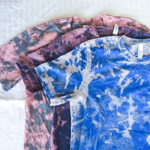 以下が含まれることがあります： ピンク、パープル、ブルーの3枚のタイダイTシャツ。シャツは重ねて置かれています。一番上のシャツは白のタイダイ模様のブルー、真ん中のシャツは黒のタイダイ模様のパープル、一番下のシャツは黒のタイダイ模様のピンクです。