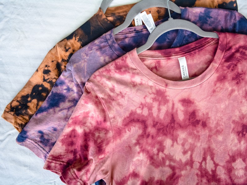 以下が含まれることがあります： 3枚の異なる色のタイダイ染めTシャツが、互いに重ねられています。一番上のシャツは、薄いピンク色で、濃いピンク色と白のタイダイ模様が施されています。真ん中のシャツは、薄い紫色で、濃い青色と白のタイダイ模様が施されています。一番下のシャツは、薄い茶色で、濃い茶色と白のタイダイ模様が施されています。シャツはすべて白い表面の上に置かれています。
