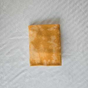 May include: A folded yellow fabric with a tie-dye pattern. The fabric has a light yellow background with darker yellow spots.