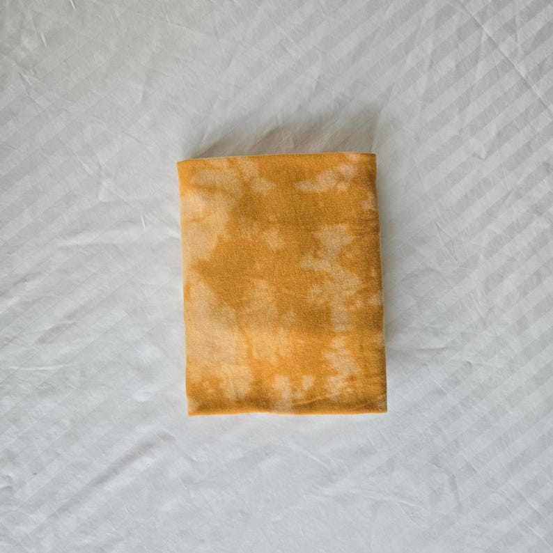 May include: A folded yellow fabric with a tie-dye pattern. The fabric has a light yellow background with darker yellow spots.
