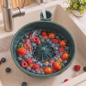 May include: A dark green ceramic colander bowl with a central cone-shaped drain feature, filled with fresh strawberries, blueberries, and cherry tomatoes being rinsed under a stream of water from a stainless steel kitchen faucet.