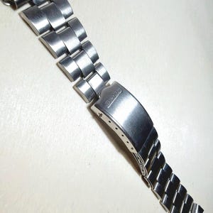 Può includere: Un cinturino per orologio in acciaio inossidabile color argento con finitura spazzolata e chiusura pieghevole. La chiusura è incisa con il marchio Seiko. Il cinturino è composto da più maglie articolate e include finali curvi per l'attacco.
