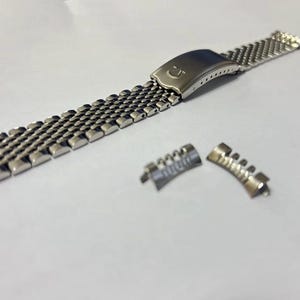 May include: A silver-toned stainless steel watch bracelet featuring a multi-link mesh design and a folding clasp with an embossed Omega logo. Two separate curved end links are positioned next to the main bracelet strap on a plain surface.