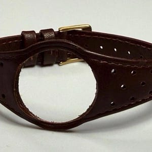 Pode incluir: Uma pulseira de relógio de couro castanho escuro com um recorte circular no centro para o mostrador do relógio. A pulseira tem um design perfurado com pequenos orifícios nas laterais e inclui uma fivela de metal dourado para prender.