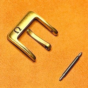 May include: A gold-toned metal watch buckle featuring the Greek letter Omega symbol engraved on the frame. The buckle consists of a square-shaped frame with a central prong and a separate silver-toned metal spring bar pin for attachment.
