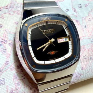 May include: A silver-tone Citizen automatic wristwatch with a black square-shaped dial, gold-tone hands, and a day-date window displaying SUN 9. The watch features a stainless steel link bracelet, a gold eagle emblem, and 21 jewels text.