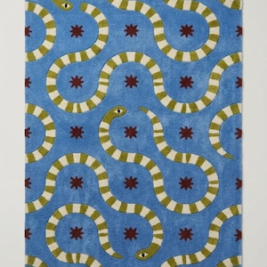 Puede incluir: Una alfombra rectangular con fondo azul brillante y un patrón repetitivo de serpientes sinuosas con rayas de color verde oliva y crema, intercaladas con pequeñas estrellas de ocho puntas de color rojo oscuro en toda la superficie.