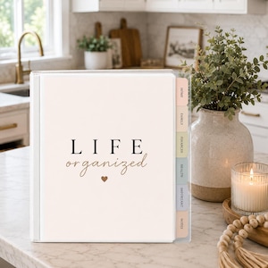 May include: A white three-ring binder with a clear front cover featuring the text LIFE organised in black and brown script with a small heart icon. The binder includes six coloured tab dividers labelled HOME, FAMILY, FINANCES, HEALTH, IMPORTANT, and NOTES.