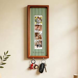 Puede incluir: Un llavero de pared rectangular de madera con un fondo de rayas verticales verdes y blancas. Muestra una tira de cuatro fotografías de una pareja e incluye el texto The Maxwell Family. Dos ganchos de metal sostienen llaveros.